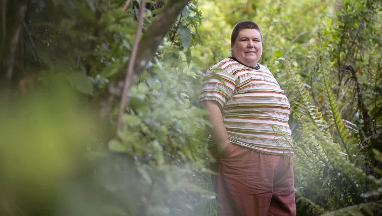 Andi Buchanan outside, surrounded by bush. They are a white non-binary person with very short brown hair, wearing orange trousers and a multi-coloured striped top. 